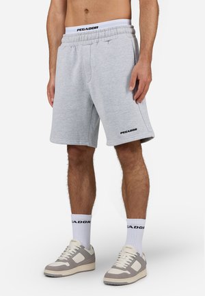 Graue Baumwoll-Sweatshorts mit einem elastischen Bund, seitlichen Taschen und schwarzem Logodetail am linken Oberschenkel, kombiniert mit hellen Sneakern und weißen Socken.