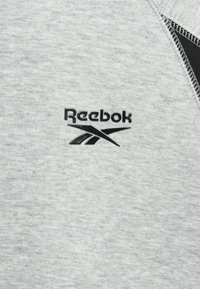 Šviesiai pilkas džemperis su tekstūruota medžiaga. Yra juodas Reebok logotipas ir juodas akcentas ant peties siūlės, pabrėžiantis sportinį dizainą.