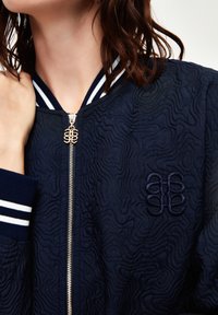 Granatowa kurtka bombowiec z teksturowanym wzorem, ściągaczami z białymi paskami i złotym zamkiem błyskawicznym z ozdobnym uchwytem.