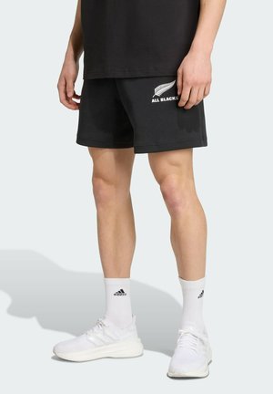 Shorts noirs en mélange de coton, ornés d'un logo "ALL BLACKS" blanc et d'un graphique de fougère. Associés à des chaussures de sport blanches et des chaussettes.