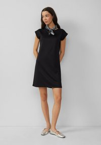 Schwarzes kurzärmliges Kleid mit rundem Ausschnitt, ausgestattet mit einer kleinen Vordertasche und einer glatten Textur. Kombiniert mit silbernen flachen Schuhen.