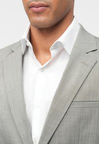 Ανοιχτό γκρίζο σακάκι με υφή, συνδυασμένο με ένα καθαρό λευκό πουκάμισο και κανονικό γιακά, που διαθέτει μία θήκη στο στήθος.