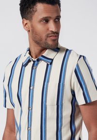 Camicia a maniche corte con bottoni, di colore crema con righe verticali blu. Presenta una singola tasca sul petto e un colletto classico. Materiale in cotone.