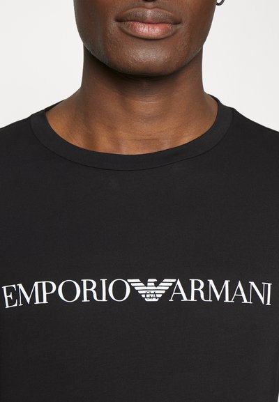 Crna pamučna majica s bijelim logotipom "EMPORIO ARMANI" i dizajnom orla. Okrugli izrez i kratki rukavi upotpunjuju dizajn.