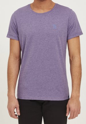Mann trägt ein schlichtes lila Kurzarm-T-Shirt mit einer kleinen gestickten blauen Palme auf der linken Brust.