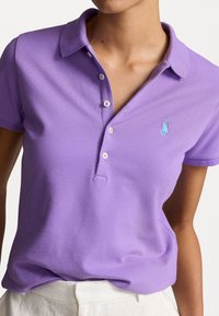 Kvinna som bär en kortärmad lavendelfärgad pikétröja med vita knappar och en liten blå broderad logotyp på bröstet, tillsammans med vita byxor.