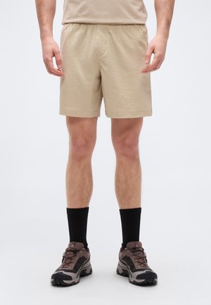 Mann steht und trägt beigefarbene, strukturierte Shorts, schwarze Socken bis zur Mitte der Wade und braune Wanderschuhe vor weißem Hintergrund.