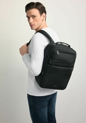 Czarny plecak wykonany z trwałej tkaniny, wyposażony w wiele zamykanych na zamek przedziałów, wyściełane paski i górny uchwyt. Prosty, prostokątny kształt.