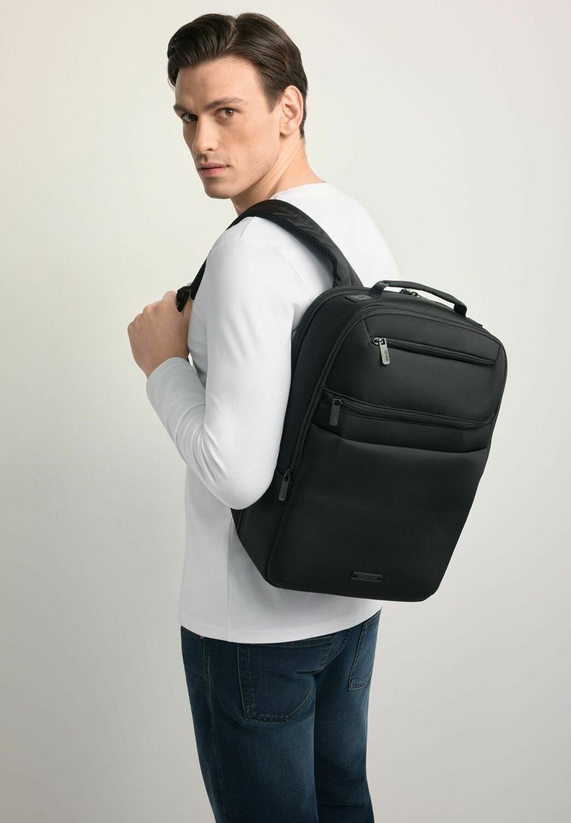 Czarny plecak wykonany z trwałej tkaniny, wyposażony w wiele zamykanych na zamek przedziałów, wyściełane paski i górny uchwyt. Prosty, prostokątny kształt.