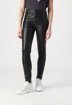Leggings neri in finta pelle a vita alta con una texture liscia e lucida, silhouette aderente, abbinati a sneakers bianche.