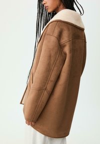 Hnedá, mäkká látková bunda s ľahkým koženým golierom, voľný strih, dlhé rukávy a prešité detaily, zobrazená z boku.