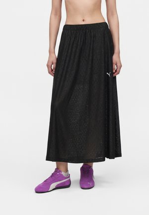Personne portant une jupe noire longueur cheville avec le logo Puma et des baskets Puma violettes, montrée de la taille aux pieds sur un fond blanc.