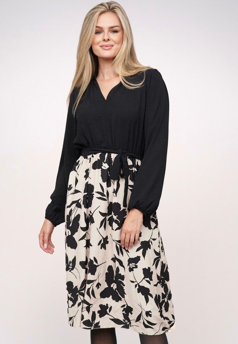 Maglietta nera a maniche lunghe con collo a V e polsini elasticizzati, abbinata a una gonna beige con un motivo floreale nero e vita con cintura.