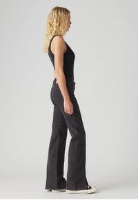 Black flared jeans with a high waist and side slits, made of denim, paired with a fitted black tank top and white sneakers.