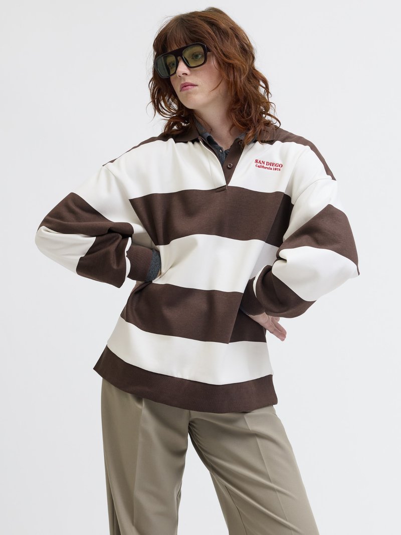 Woman wearing oversized brown and white striped rugby shirt with "San Diego California 1975" text, beige pants, and large dark sunglasses.
