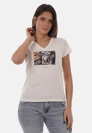 Femme aux cheveux bruns raides portant un t-shirt blanc à col en V avec un motif graphique et un jean gris clair, posant avec une main près du visage.