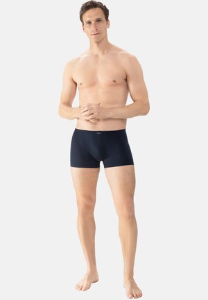 Homme debout pieds nus portant un boxer bleu marine foncé, les mains jointes et une expression neutre sur un fond uni.
