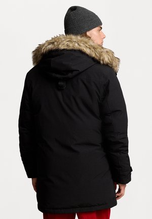 Parka noire isolée avec une capuche doublée en fausse fourrure, dotée de manches longues, d'un cordon de serrage et d'un design épuré et matelassé. Bonnet en tricot gris porté.