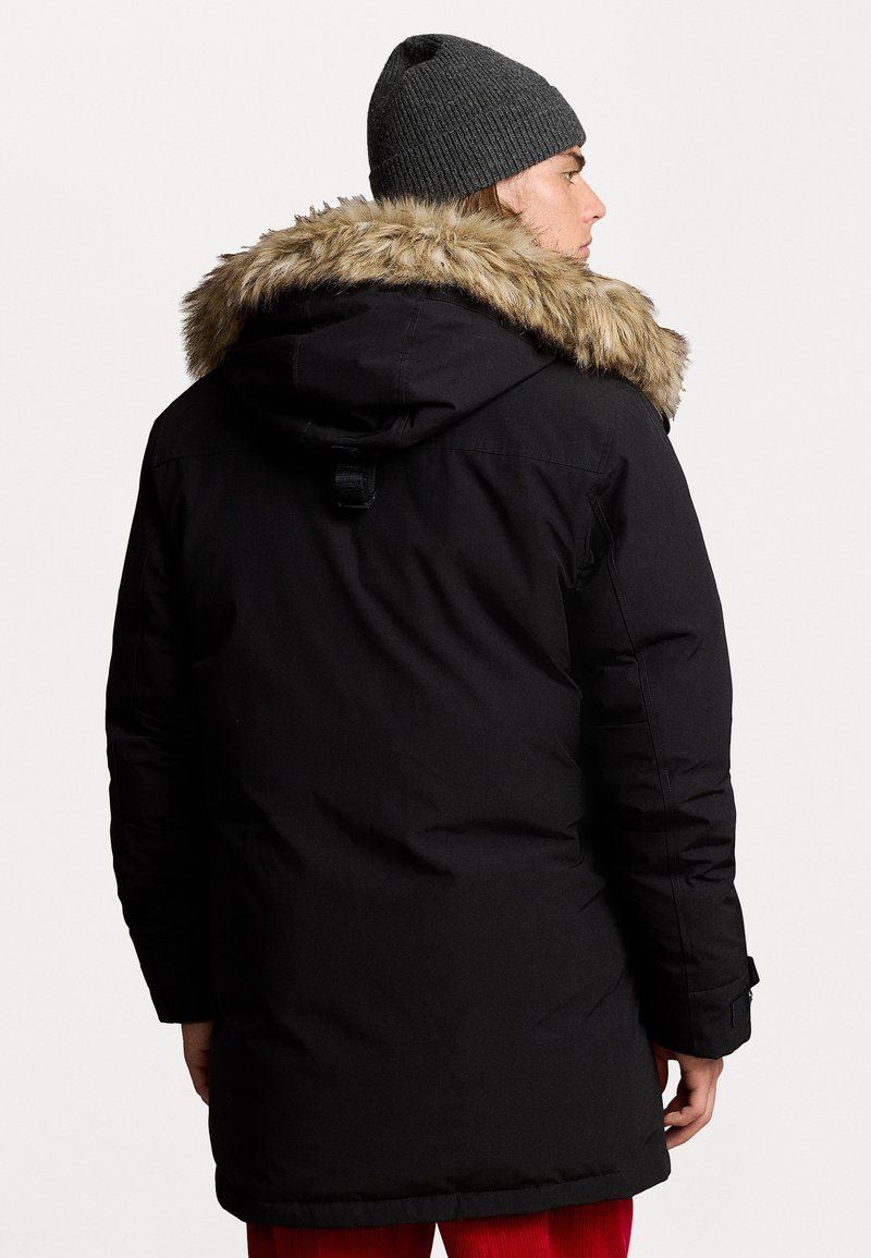 Parka nera isolata con cappuccio foderato in pelliccia sintetica, dotata di maniche lunghe, coulisse e un design elegante e imbottito. Indossato un berretto grigio a maglia.