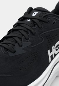 Chaussure de sport noire avec un dessus texturé, dotée de lacets ton sur ton et d'un logo blanc. La semelle intermédiaire possède un design en relief visible et un bord contrastant.