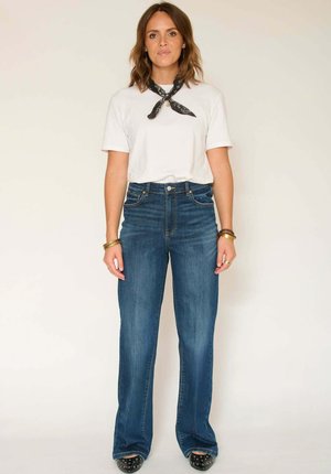 T-shirt blanc à manches courtes associé à un jean bleu taille haute à jambes larges. Accessoires : foulard bandana noir et bracelets. Chaussures noires cloutées.