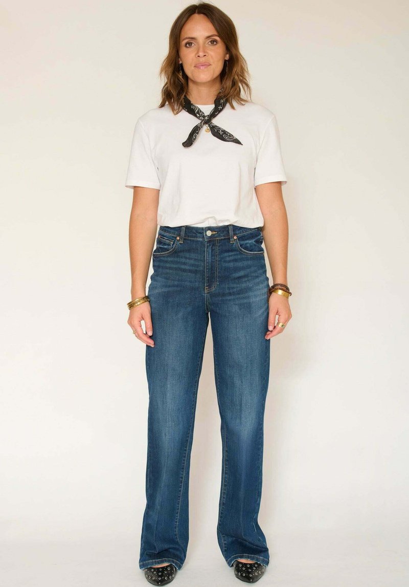 T-shirt blanc à manches courtes associé à un jean bleu taille haute à jambes larges. Accessoires : foulard bandana noir et bracelets. Chaussures noires cloutées.