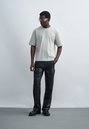 Man draagt een grijs oversized T-shirt, zwarte leren cargobroek, zwarte schoenen en transparante oversized zonnebril, staand tegen een effen achtergrond.