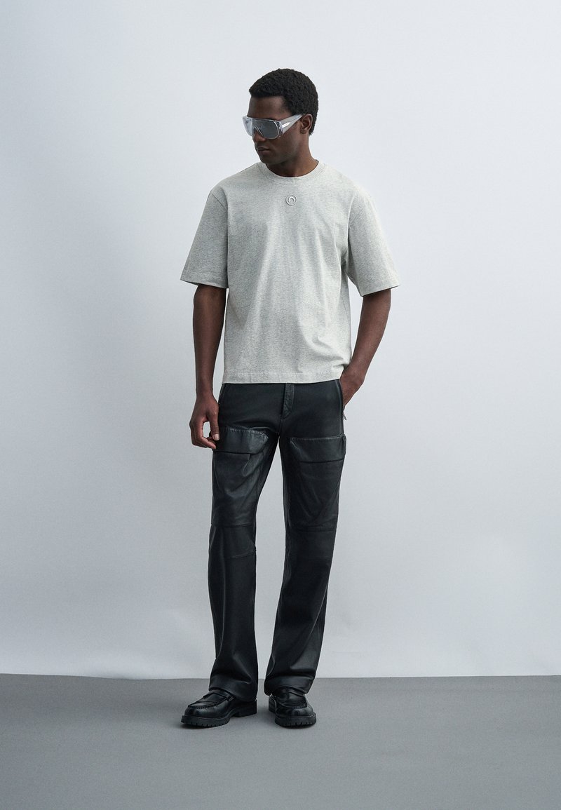 Άνδρας που φορά γκρι μεγάλο μπλουζάκι, μαύρο δερμάτινο cargo παντελόνι, μαύρα παπούτσια και διάφανα oversized γυαλιά ηλίου, στέκεται μπροστά σε απλό φόντο.