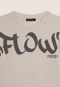 Camiseta de algodón color crema con gran texto negro que dice "FLOW" y "FREDDY". Cuello redondeado y mangas cortas. Textura suave.