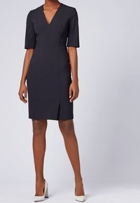 Mörkblå klänning med V-ringning, korta ärmar och en slits framtill. Tillverkad av ett mjukt tyg med en figurnära silhuett, kombinerad med vita klackar.