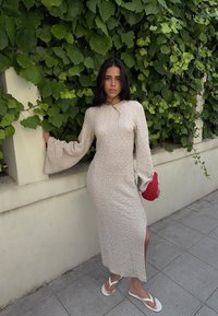 Kreemjas, tekstuuriga pikad varrukad kleit, külje lõhega, koos valgete plätudega ja punase heegeldatud kotiga, rohelise lehtede taustal.