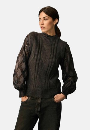 Un pull en maille noir avec des motifs en câble texturés. Des manches longues et transparentes, à motifs, apportent un contraste. Encolure et ourlet côtelés. Porté avec un jean foncé.
