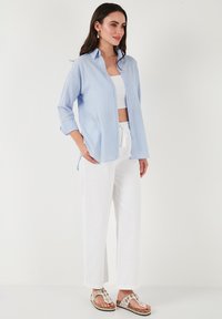 Light blue button-up shirt with vertical stripes, rolled sleeves, worn over a white crop top and paired with white pants and decorative sandals.