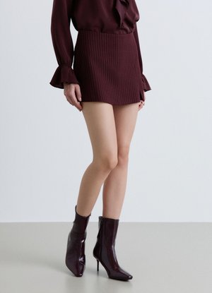 Donna che indossa una minigonna bordeaux a righe sottili, una camicetta abbinata con polsini arricciati e stivaletti lucidi bordeaux scuro con tacco alto.