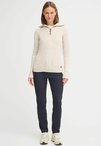 Cable-knit cream sweater with a high collar, zip closure, and ribbed cuffs, paired with navy slim-fit pants and light-colored sneakers.