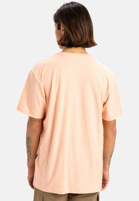 Camiseta de manga corta en melocotón claro con un ajuste relajado, textura suave, escote redondo y una etiqueta lateral discreta cerca del dobladillo.