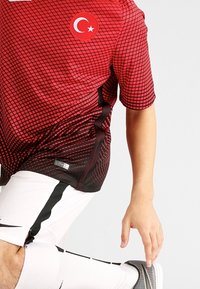 Maillot de football en maille rouge et noir avec emblème du drapeau turc, doté de manches courtes et d'un motif en grille. Short blanc avec accents noirs.