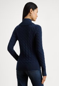 Moteris, stovinti šonu, rodanti tamsiai mėlyno kabelinio mezgimo megztinio nugarą, su ribėtu aukštu apykakliu ir rankogaliais, dėvinti tamsiai mėlynas džinsines kelnes.