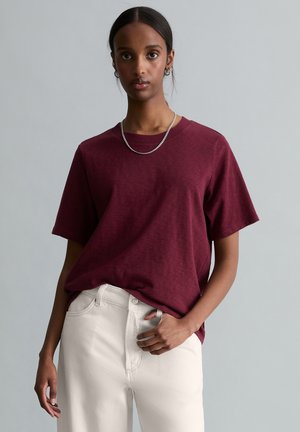 Person wearing a burgundy short-sleeve shirt, white pants, silver chain necklace, and small hoop earrings against a plain gray background.