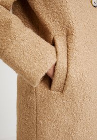 Beige textured coat made of fuzzy material with a pocket at the hip and a visible button near the collar.