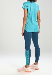 Turkos kortärmad träningsskjorta med en slits bak, tillsammans med marinblå leggings med diagonala randiga mönster och reflekterande detaljer.