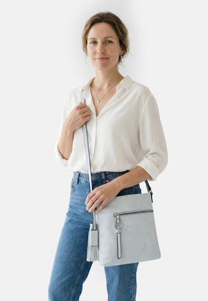 Woman with light brown hair wearing a white blouse and blue jeans holds a light grey crossbody bag with a front zipper pocket.