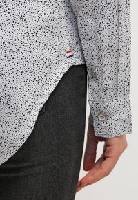 Biała koszula z czarnym, nakrapianym wzorem, mankiety zapinane na guziki oraz mała naszywka z logo w kolorach czerwonym, białym i niebieskim blisko dolnego brzegu. Noszona z ciemnoszarymi spodniami.