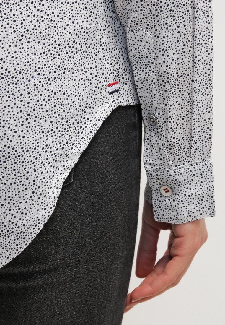 Biała koszula z czarnym, nakrapianym wzorem, mankiety zapinane na guziki oraz mała naszywka z logo w kolorach czerwonym, białym i niebieskim blisko dolnego brzegu. Noszona z ciemnoszarymi spodniami.