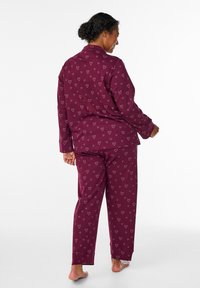 Tummanpunainen sydänkuviollinen pyjamapuku, jossa on pitkähihainen paita ja siihen sopivat housut, valmistettu pehmeästä kankaasta ja väljässä istuvuudessa.