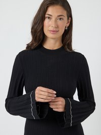 Mulher com cabelo castanho de comprimento médio, vestindo um suéter preto com textura canelada, mangas largas e detalhes brancos, com as mãos unidas à altura da cintura.
