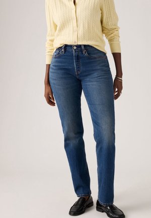 Personne portant un pull jaune clair boutonné, un jean droit bleu et des mocassins noirs, debout devant un fond uni.