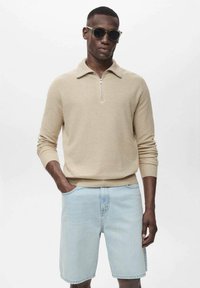 Suéter de punto beige con un cuello con cremallera, combinado con pantalones cortos de mezclilla azul claro. El modelo lleva gafas de sol oscuras y está de pie frente a un fondo liso.