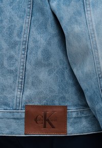 Světlá modrá džínová bunda s výrazným prošíváním a hnědou koženou nášivkou s vyraženým nápisem "cK Calvin Klein" na spodní části zad.