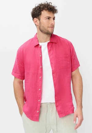 Mann trägt ein leuchtend pinkes kurzärmeliges Hemd über einem weißen T-Shirt mit hellen Hosen, Hände in den Taschen, schaut nach rechts.